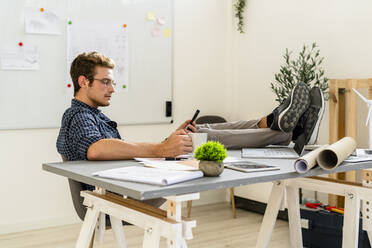 Young man with legs on desk using mobile phone at office - GIOF08894