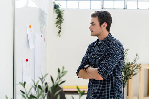 Mann mit verschränkten Armen, der im Büro stehend auf ein Whiteboard schaut - GIOF08902