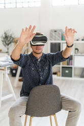 Young man with arms raised using visual reality simulator while sitting on chair at office - GIOF08917