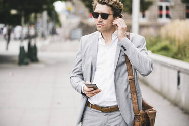 Businessman connecting in-ear headphones with mobile phone while standing at sidewalk in city - UUF21561