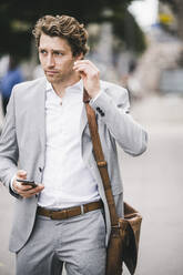 Businessman with mobile phone wearing in-ear headphones while standing in city - UUF21562
