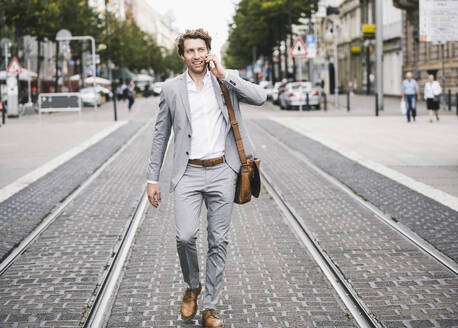 Smiling man talking on mobile phone while walking at tramway in city - UUF21570