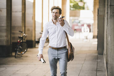 Lächelnder Mann, der am Telefon spricht, während er an einem Gebäude in der Stadt spazieren geht - UUF21581