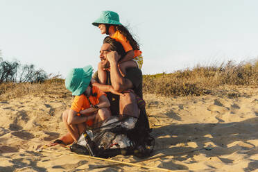 Vater und Töchter sitzen auf Sand mit Müllsack während eines sonnigen Tages - ERRF04471