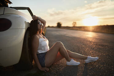 Young woman sitting on road against car during sunset - ZEDF03821