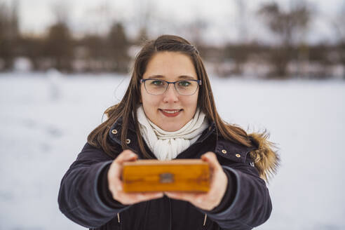 Nahaufnahme einer lächelnden jungen Frau, die ein Weihnachtsgeschenk hält, während sie im Winter im Freien steht - JSCF00160