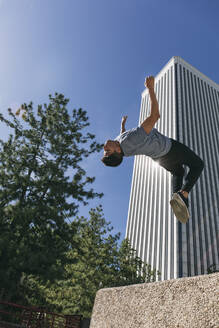 Young man jumping over wall performing parkour against clear blue sky in city - ABZF03326