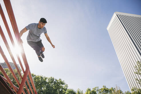 Young man jumping over railing performing parkour against clear sky in city - ABZF03328