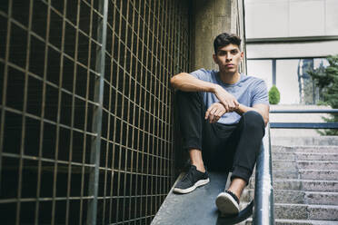 Serious young man sitting on retaining wall by fence - ABZF03336