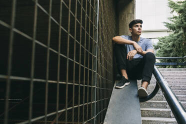 Thoughtful young man sitting on retaining wall by fence - ABZF03337