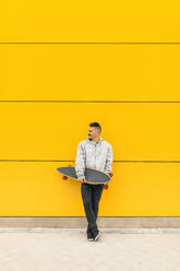 Porträt eines jungen Mannes, der mit einem Skateboard in der Hand an einer gelben Wand lehnt - MRRF00480