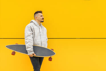 Porträt eines jungen Mannes, der mit einem Skateboard in der Hand an einer gelben Wand lehnt - MRRF00481