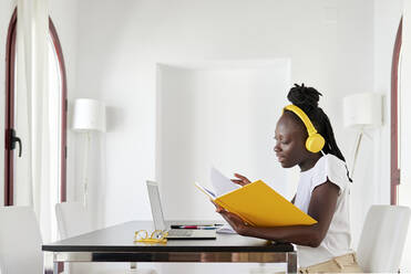 Businesswoman wearing headphones reading book at desk in home office - KIJF03267