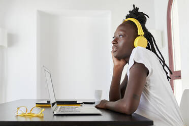 Nachdenkliche Geschäftsfrau mit Laptop auf dem Schreibtisch und Kopfhörern, während sie zu Hause sitzt - KIJF03270
