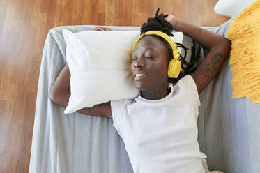 Young woman with eyes closed listening music over headphones while lying on sofa in living room - KIJF03276