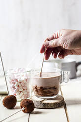 Hand einer Frau, die heiße Schokolade mit Trüffellutschern und Marshmallows auf einem Tisch zu Hause mischt - SBDF04387