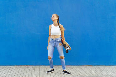 Cheerful woman holding skateboard standing on footpath against blue wall - PGF00036