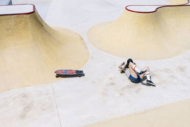 Junge Frau mit Skateboard auf Sportrampe liegend an einem sonnigen Tag - PGF00056