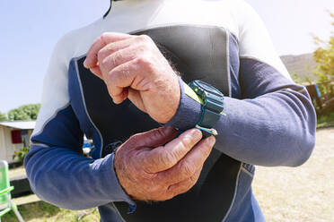 Close-up of senior man wearing wetsuit adjusting wristwatch at beach during sunny day - PGF00058