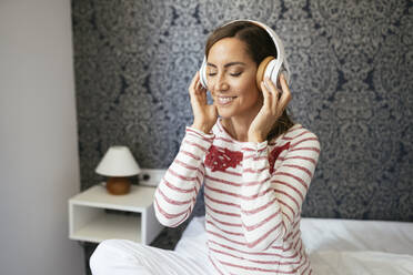 Lächelnde Frau mit geschlossenen Augen hört Musik, während sie zu Hause auf dem Bett sitzt - JSMF01712