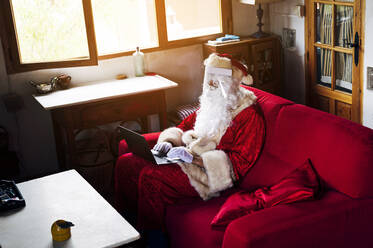 Man wearing Santa Claus costume using laptop while sitting on sofa at home - PGF00062