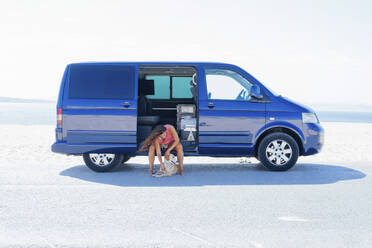 Woman petting dog while sitting on blue motor home at beach against clear sky - PGF00071