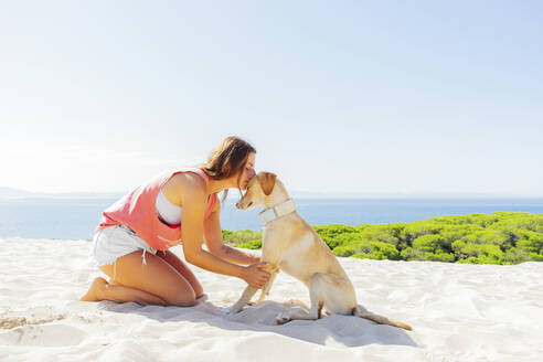 Frau küsst Hund, während kniend am Strand gegen klaren Himmel während des sonnigen Tages - PGF00073