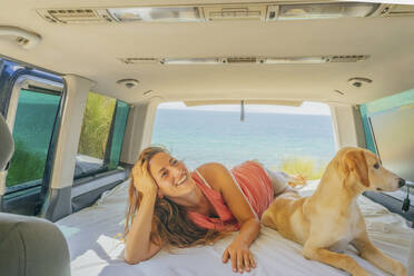 Lächelnde erwachsene Frau mit Hund, die sich auf dem Bett eines Wohnmobils am Meer entspannt - PGF00076