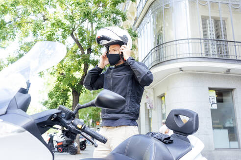 Männlicher Motorradfahrer setzt seinen Helm auf, während er neben einem Motorroller steht - PGF00081