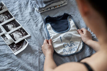 Pregnant woman folding baby clothes by ultrasound on bed at home - BZF00584