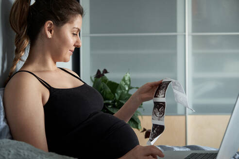 Schwangere Frau hält Ultraschall-Scans, während sie einen Laptop auf dem Bett zu Hause benutzt - BZF00585
