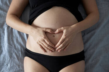 Schwangere Frau macht Herzform auf dem Bauch, während auf dem Bett zu Hause liegen - BZF00586