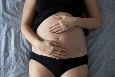 Schwangere Frau berührt den Bauch, während sie zu Hause auf dem Bett liegt - BZF00587