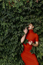 Young woman wearing red dress leaning on green leaves - TCEF01135