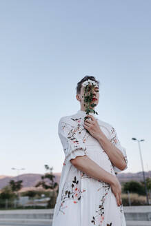 Gender fluid man covering face with bouquet of daisies while standing on street during sunset - TCEF01149