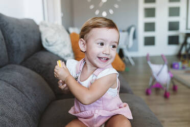 Baby girl holding biscuit while sitting on sofa at home - EBBF00772