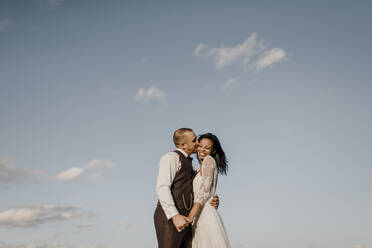 Bridegroom kissing bride on cheek during sunny day - SMSF00338