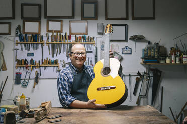 Lächelnder Mann, der eine Gitarre hält, während er an einer Werkbank steht - DGOF01485