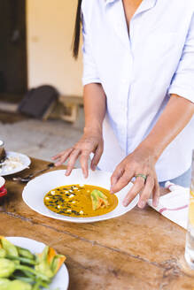 Frau garniert Suppe mit Gemüse, während sie am Tisch im Hinterhof steht - FMOF01075