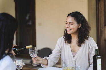 Woman pouring champagne in friend's wineglass while sitting by table at back yard - FMOF01077
