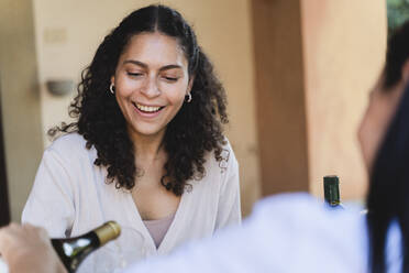Friend serving pouring champagne in woman glass while sitting at back yard - FMOF01078