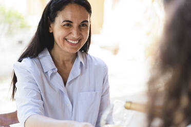 Friend smiling while looking at woman pouring wine in glass at back yard - FMOF01082