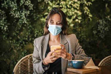 Woman text messaging on smart phone wearing protective face mask while sitting in cafe - GRCF00368