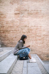 Young woman using smart phone while sitting on staircase in city - GRCF00385