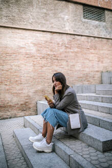 Junge Frau benutzt ihr Smartphone mit der Hand am Kinn, während sie auf einer Treppe in der Stadt sitzt - GRCF00386