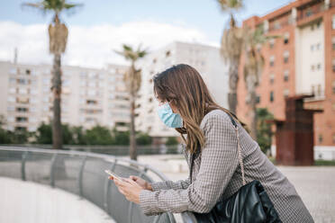 Young woman wearing face mask while using smart phone leaning on railing in city - GRCF00394