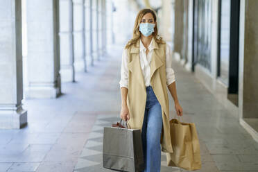 Frau mit Gesichtsmaske, die eine Tasche trägt, während sie in der Lobby eines Geschäfts in der Stadt spazieren geht - JSMF01723