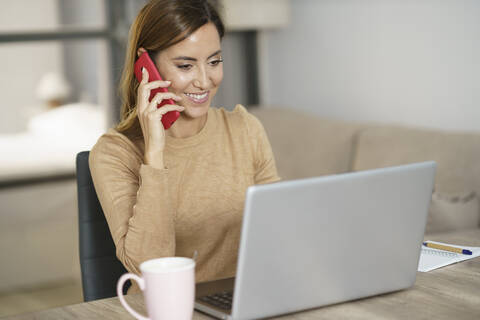 Smiling businesswoman talking on phone while working on laptop at home stock photo