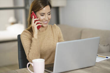 Smiling businesswoman talking on phone while working on laptop at home - JSMF01741