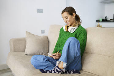 Smiling woman writing on book while sitting on sofa at home - JSMF01743